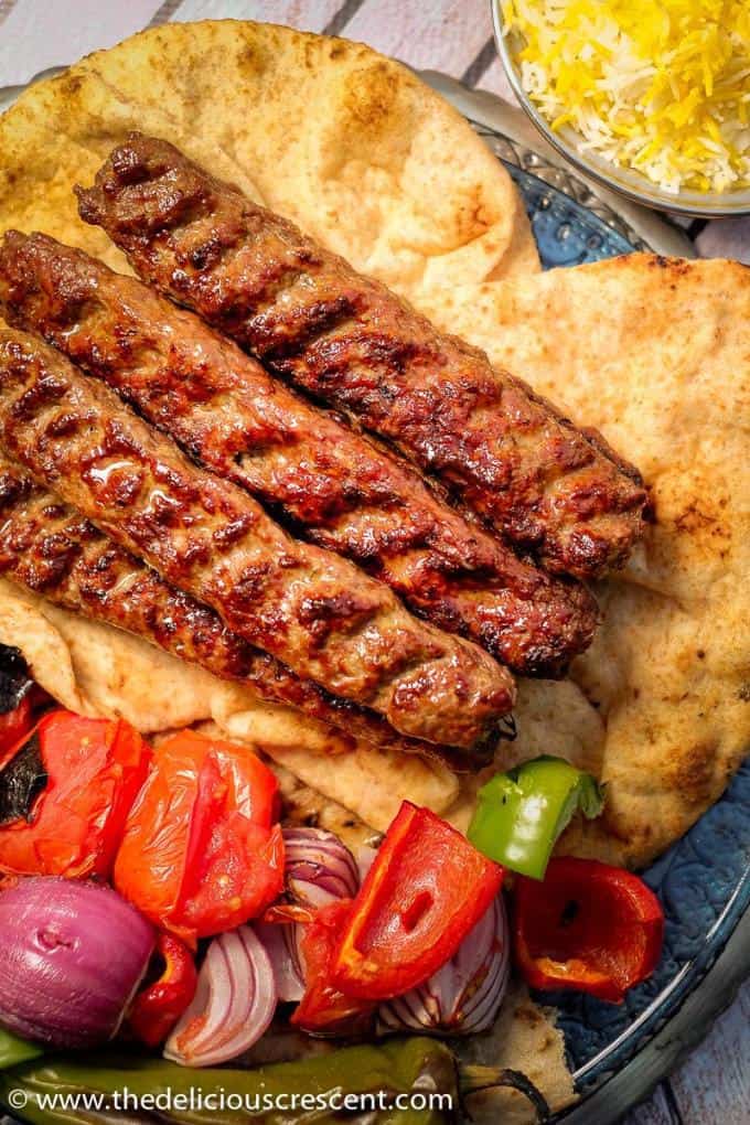 Overhead view of koobideh kabob skewers with vegetables all placed on flat breads. 